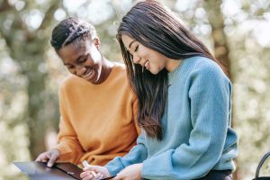 Two women sit outside looking down and writing