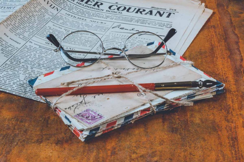 A package of letters bound with string on the table next to a newspaper. On top a pair of vintage glasses.