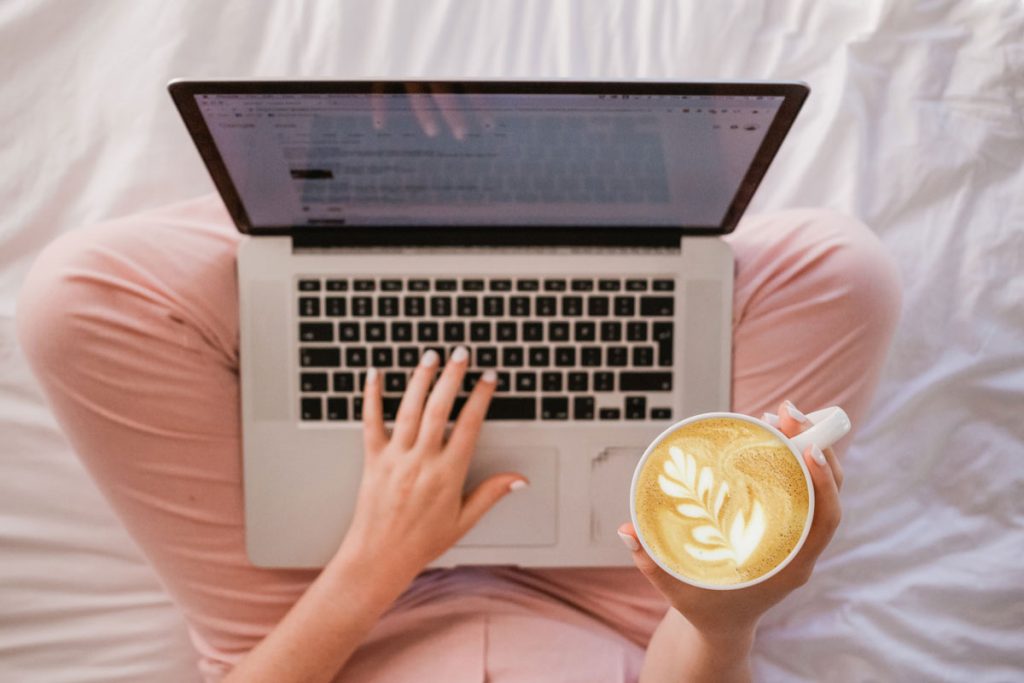 Half a photo of someone sitting on the bed with pink trousers holding a laptop and a coffee
