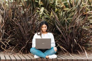 A woman sits on a wooden dock with a laptop surrounded by reeds while registering a postcard from PenPal