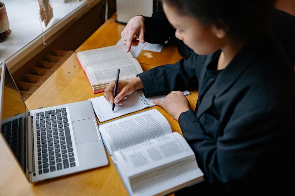 Someone writing a note with a laptop and dictionary beside her.