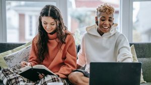 Two women sit learning a new language, one on the laptop and the other with a book